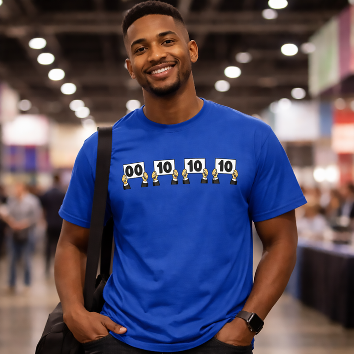 Male model wearing a royal blue “Everything is Judged in Binary” short-sleeve unisex t-shirt at a geek convention, featuring judges holding scorecards reading 00 10 10 10 in a Hitchhiker-inspired binary joke design.