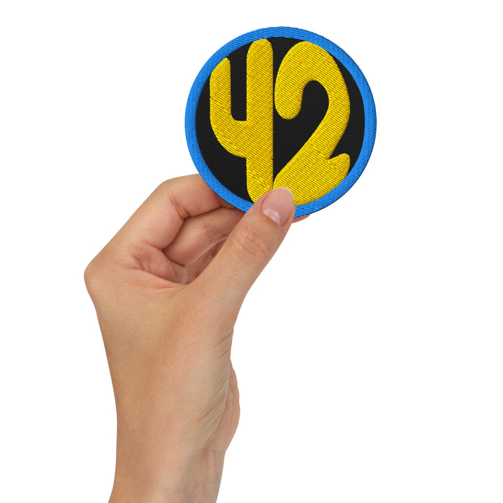 Hand holding a 3-inch embroidered 42 circle patch with gold numbers on a black background and blue border.