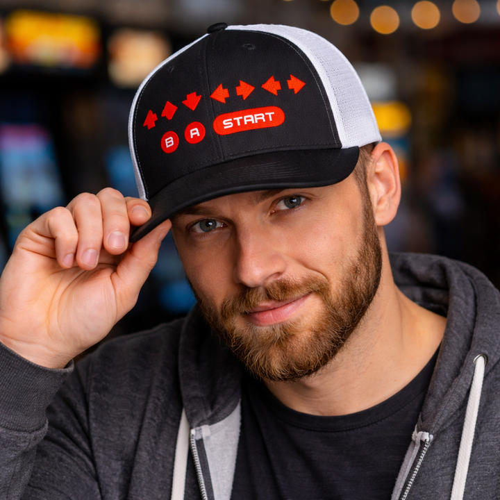 Model wearing a black and white mesh-back trucker cap embroidered with a retro video game cheat code design featuring arrow icons and B A Start buttons