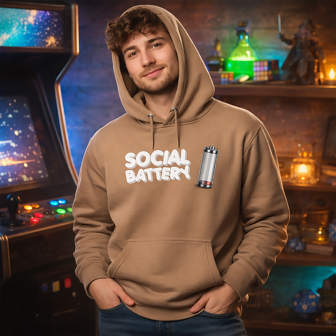 Model wearing the latte Social Battery hoodie in a cozy geek-themed room with retro arcade vibes, warm ambient lighting, and fantasy-inspired shelf décor.