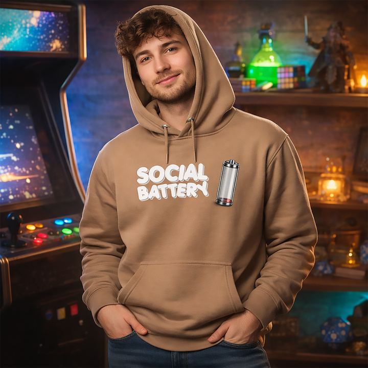 Model wearing the latte Social Battery hoodie in a cozy geek-themed room with retro arcade vibes, warm ambient lighting, and fantasy-inspired shelf décor.
