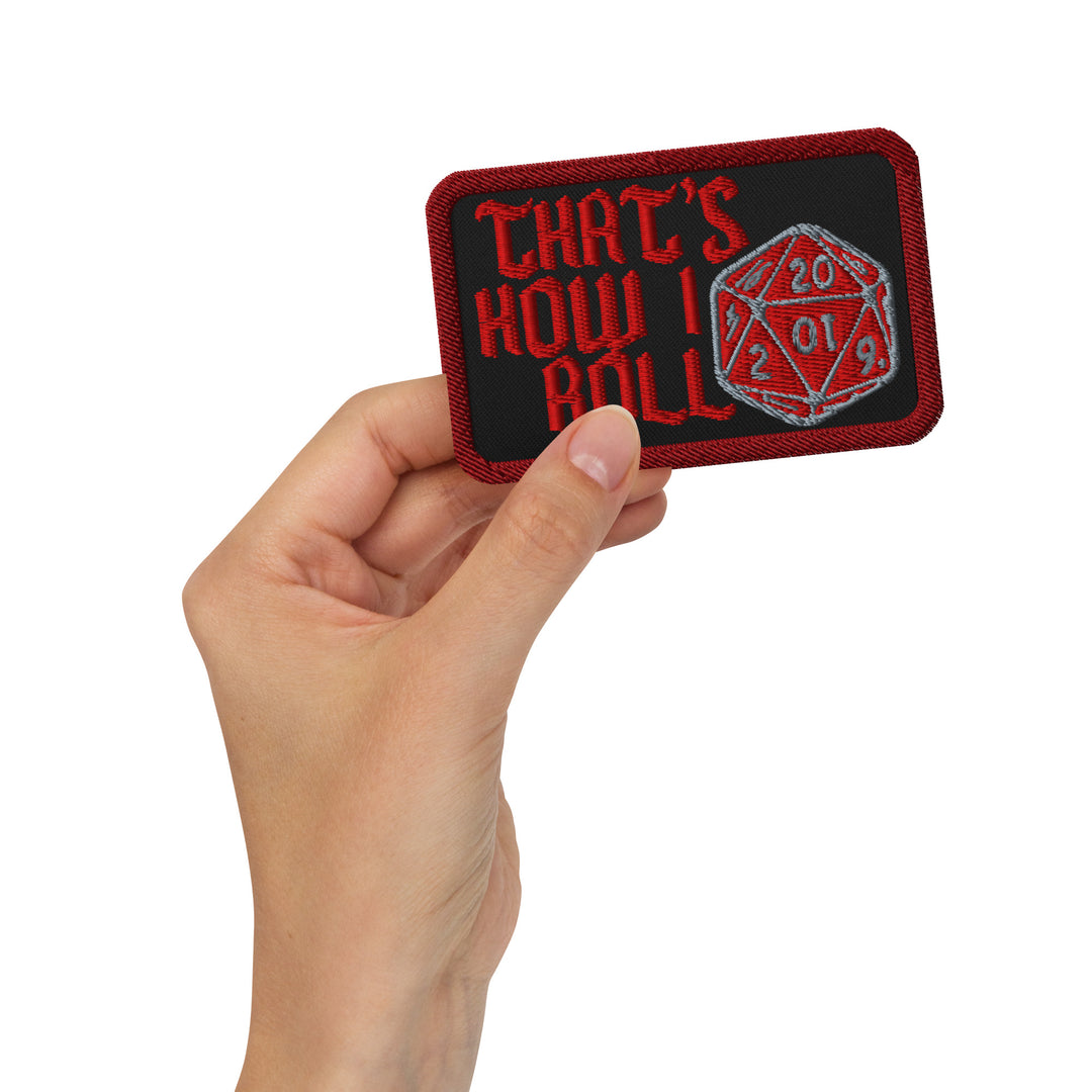 Hand holding the black “That’s How I Roll” embroidered patch with red lettering and a red-and-silver D20 design.