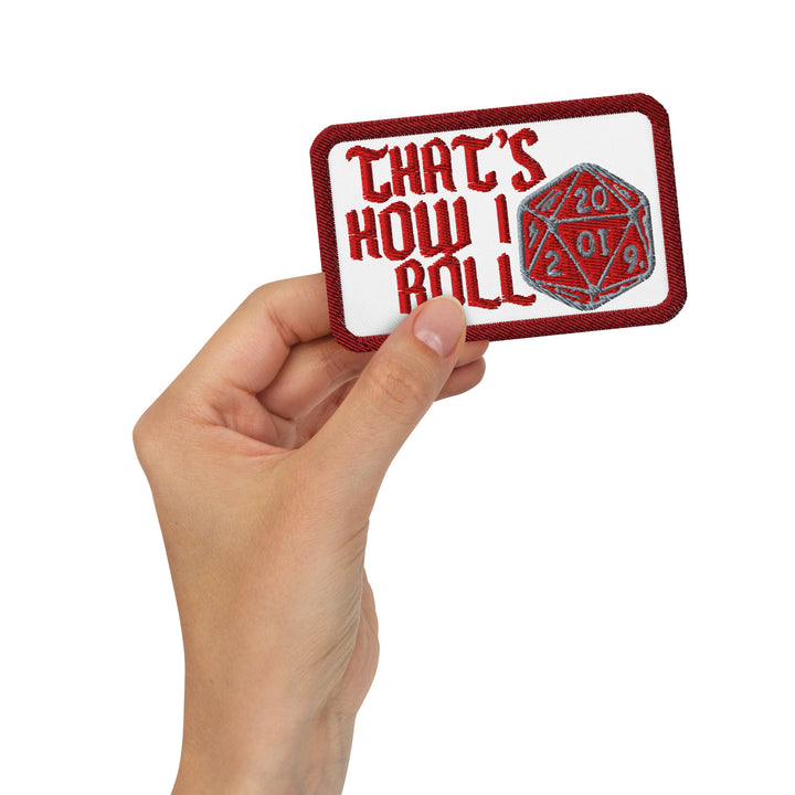 Hand holding the white “That’s How I Roll” embroidered patch with red lettering and a red-and-silver D20 design.