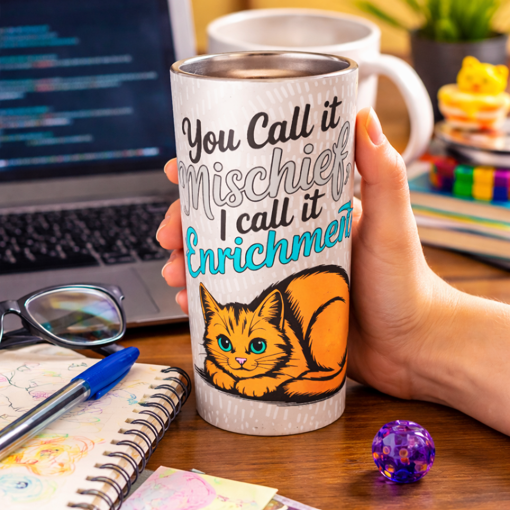Hand holding a white 20 oz tapered stainless steel tumbler with “You Call it Mischief, I Call it Enrichment” orange cat design on a cluttered geek desk with laptop, notebook, and glasses.